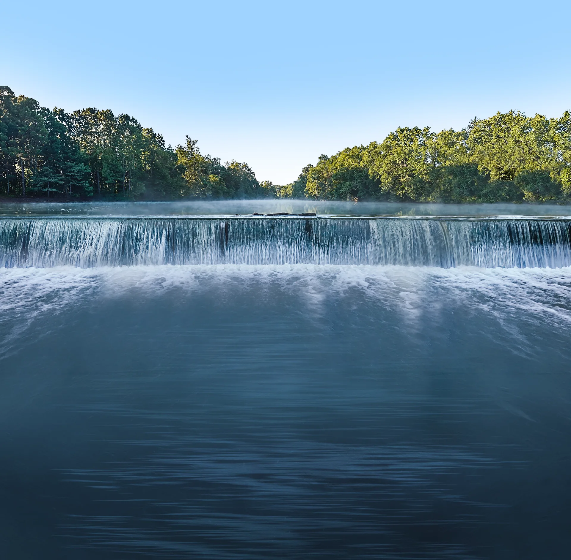 Low-head dam waterfall on a misty tree-lined river